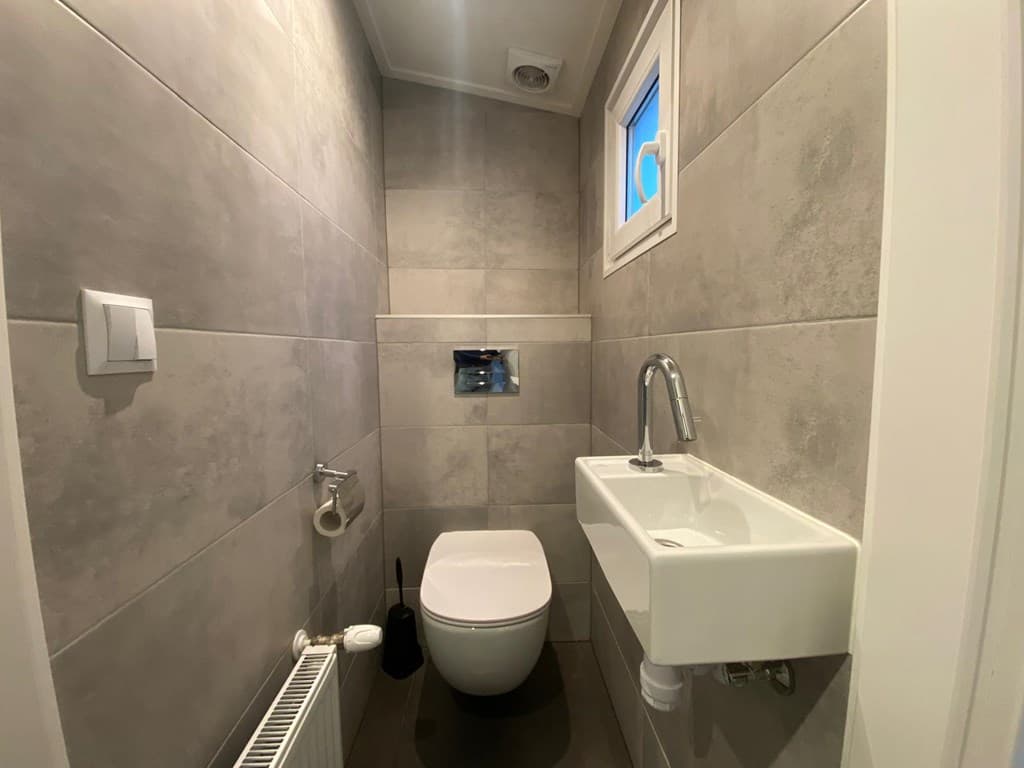 Modern toilet met grijze tegelwanden, wandtoilet en fonteintje bij het raam