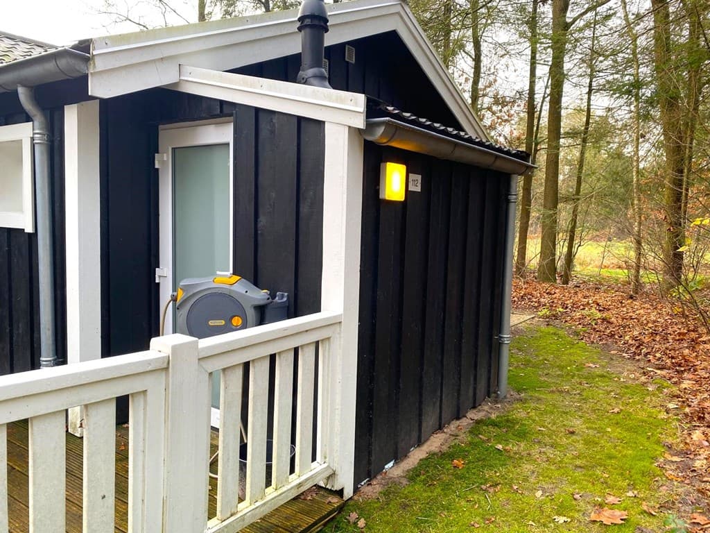 Zijaanzicht van een houten woning met een balkon en een wateraansluiting, omringd door bomen en gevallen bladeren