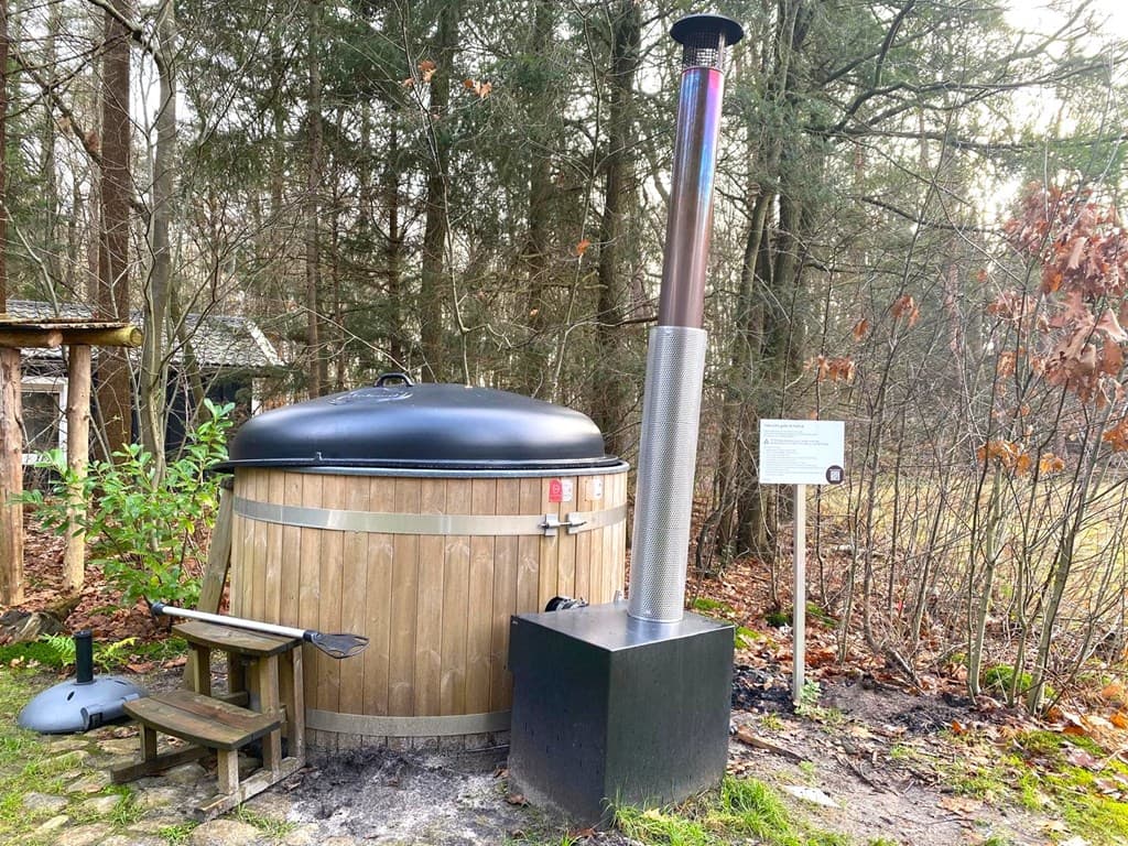 Houten hottub met een metalen kachel in een bosachtige omgeving met een bankje ernaast.