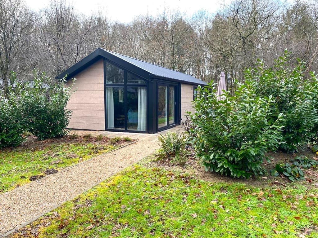 Moderne vakantiewoning met grote ramen, omgeven door groen en een grindpad naar de ingang.