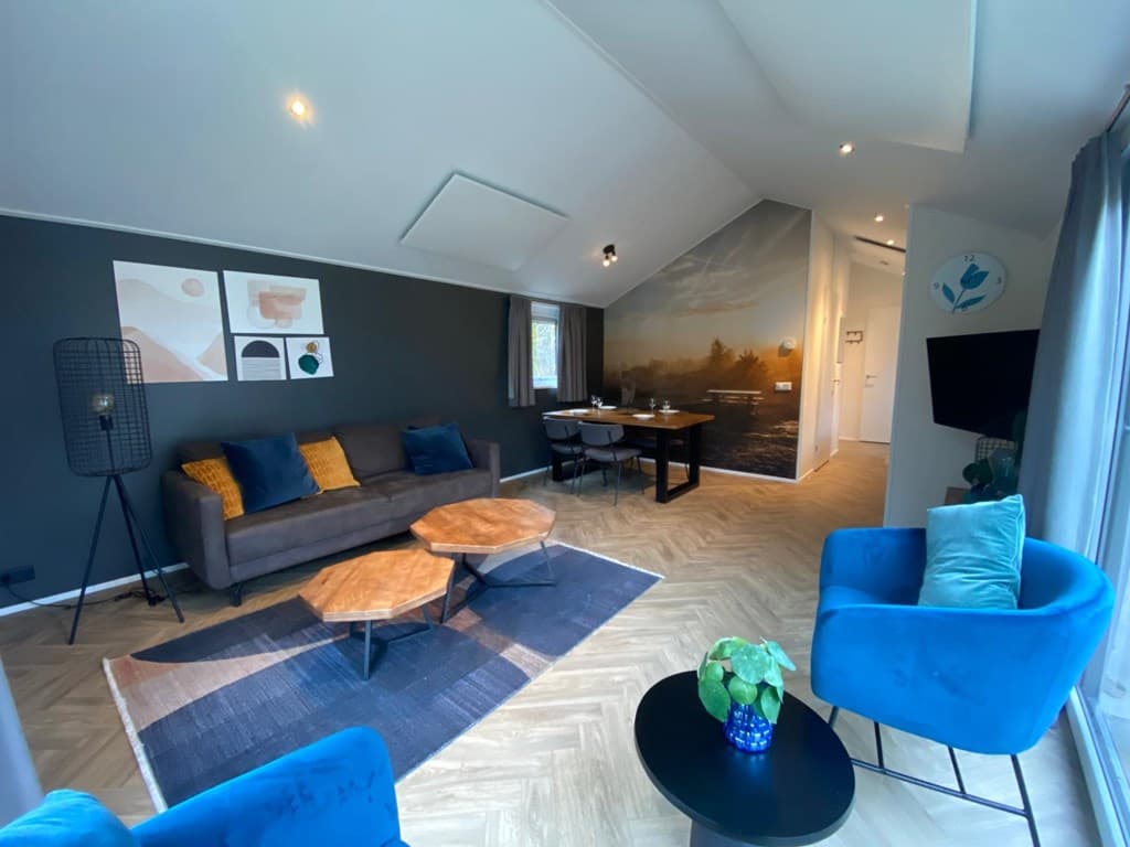 Woonkamer met een grijze bank, blauwe fauteuils, houten tafels en een eettafel met stoelen in een moderne inrichting