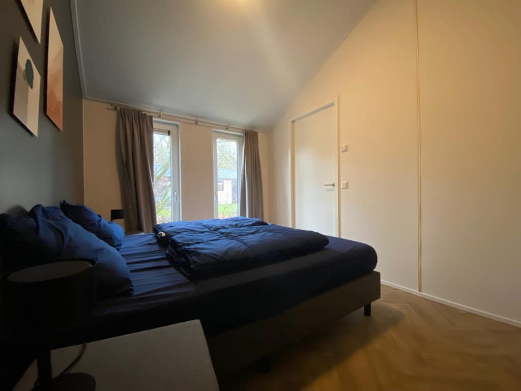 Slaapkamer met een bed met blauwe dekbedden, ramen met gordijnen en een deur naar buiten.