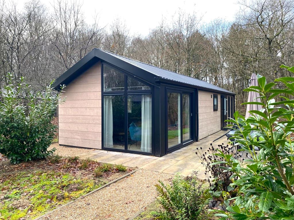 Moderne vakantiewoning met grote ramen, ligt omgeven door bomen en een tuin met planten en een terras.