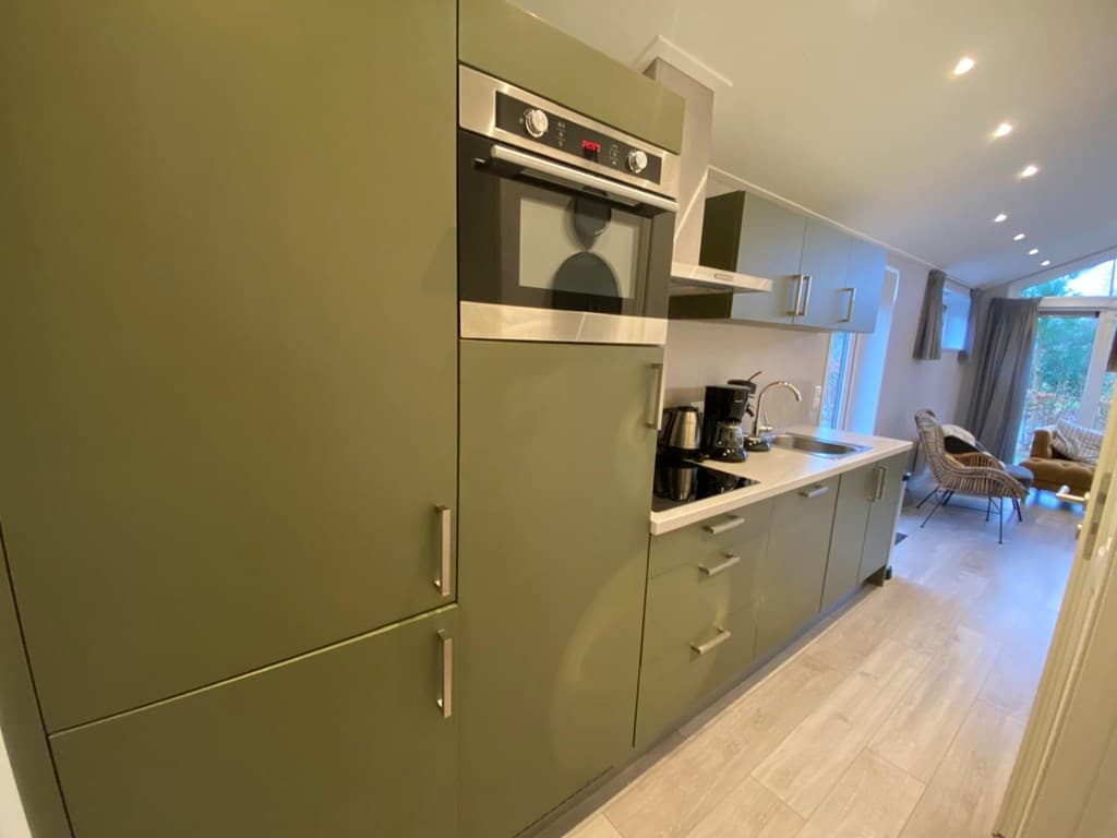 Keuken met groene kasten, oven, koffiezetapparaat en uitzicht op buiten via grote ramen.