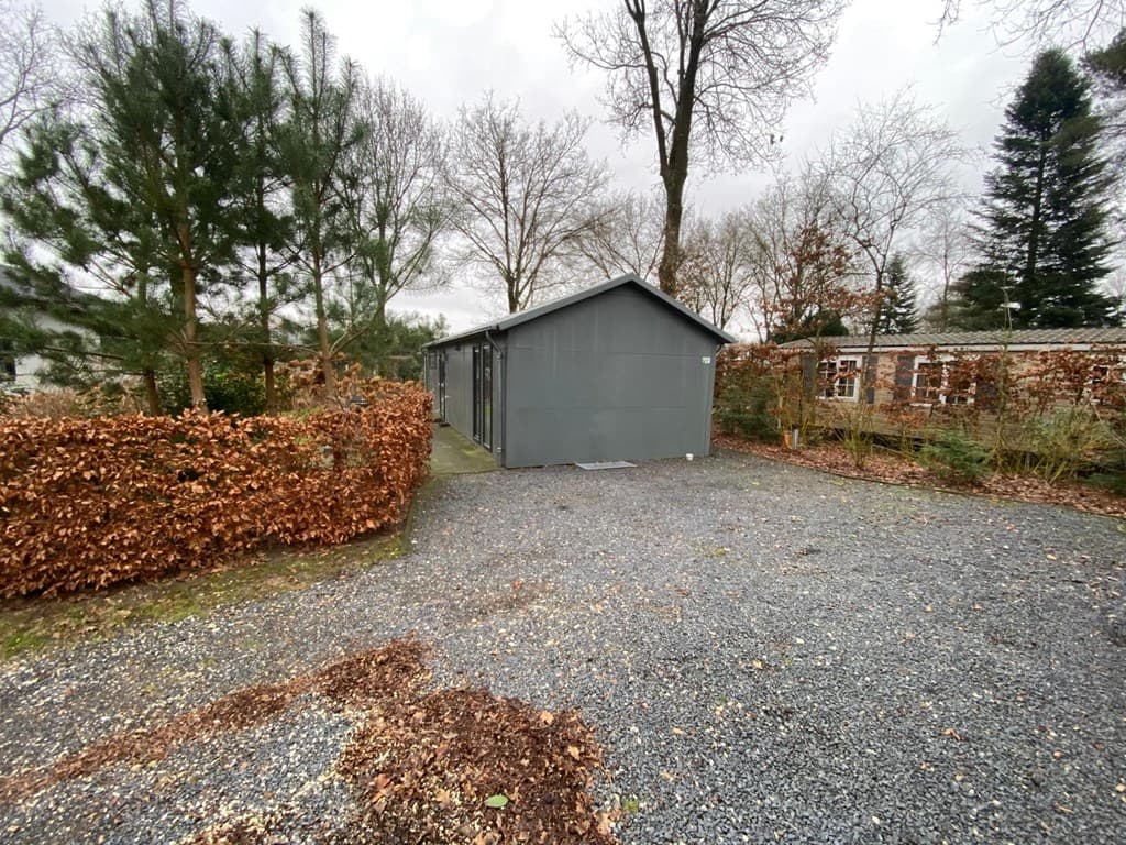 Grijze vakantiewoning omringd door begroeiing en grindpad, met bomen en struiken op de achtergrond