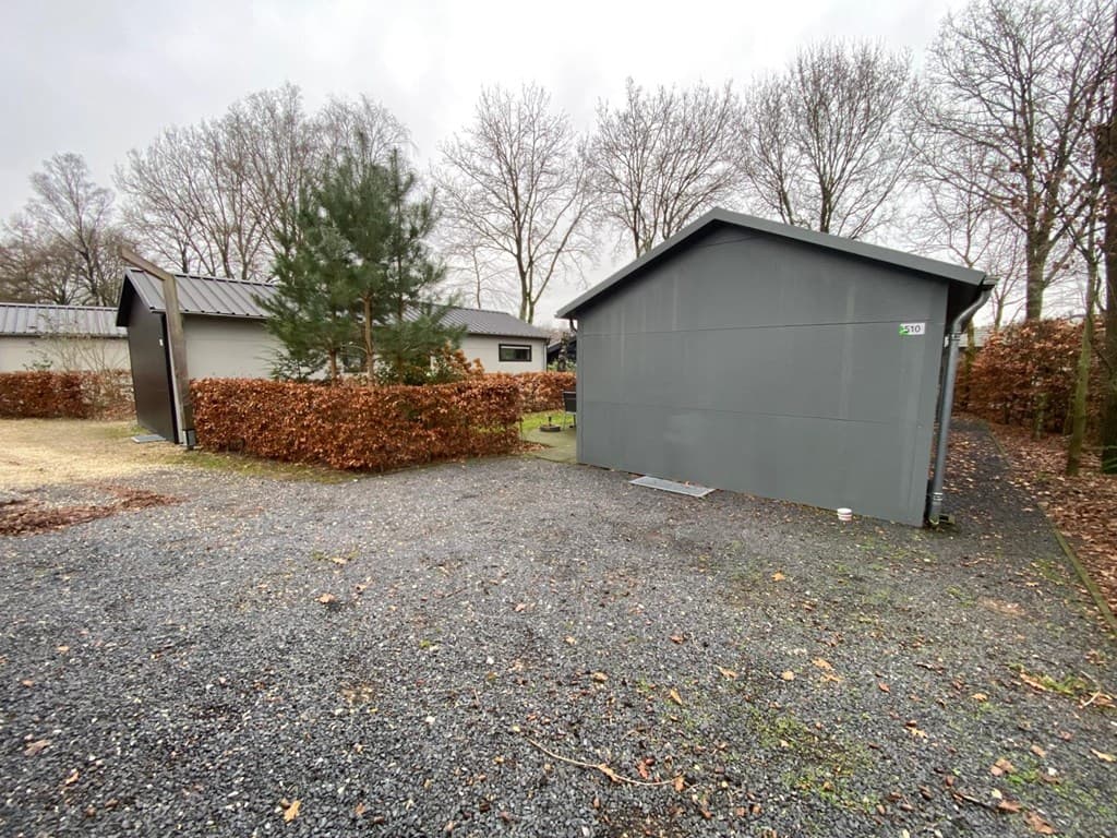 Grijze vakantiewoning met een naastgelegen schuur, omgeven door een gravelpad en groene hagen in een bosrijke omgeving.