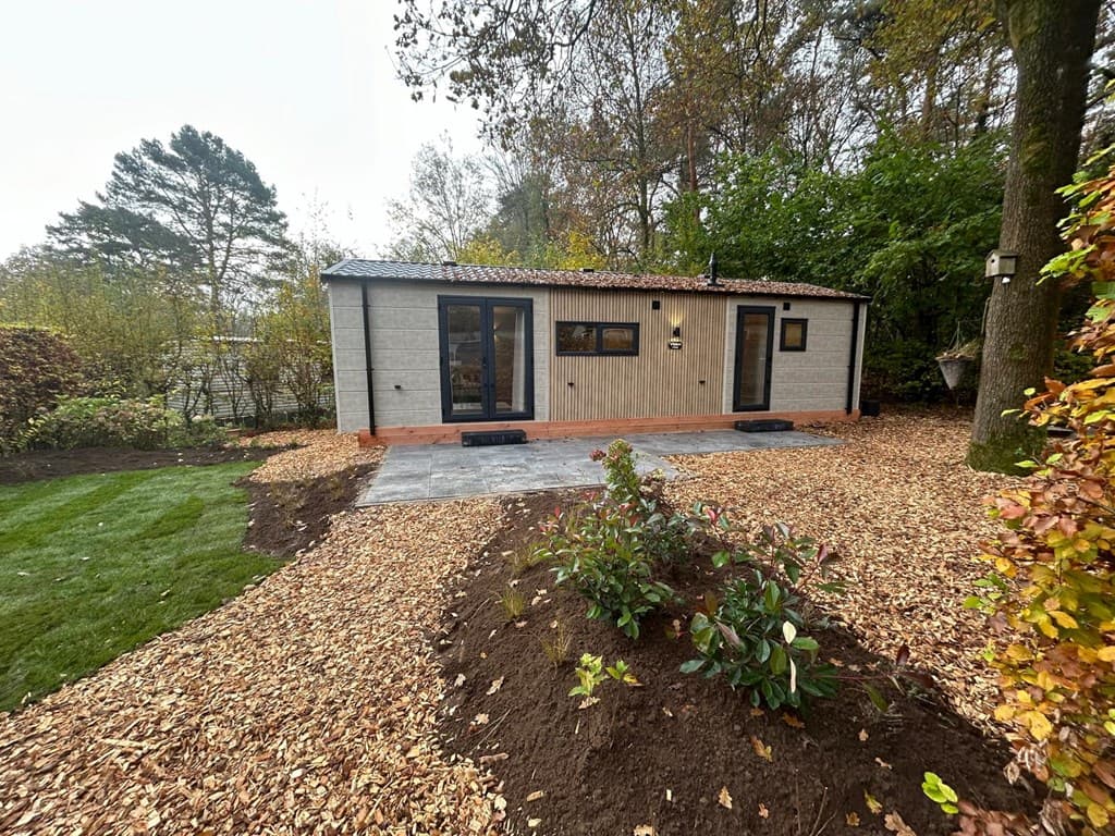 Moderne vakantiewoning met houten gevel, grote ramen en omringd door bomen en een aangelegde tuin met houtsnippers.