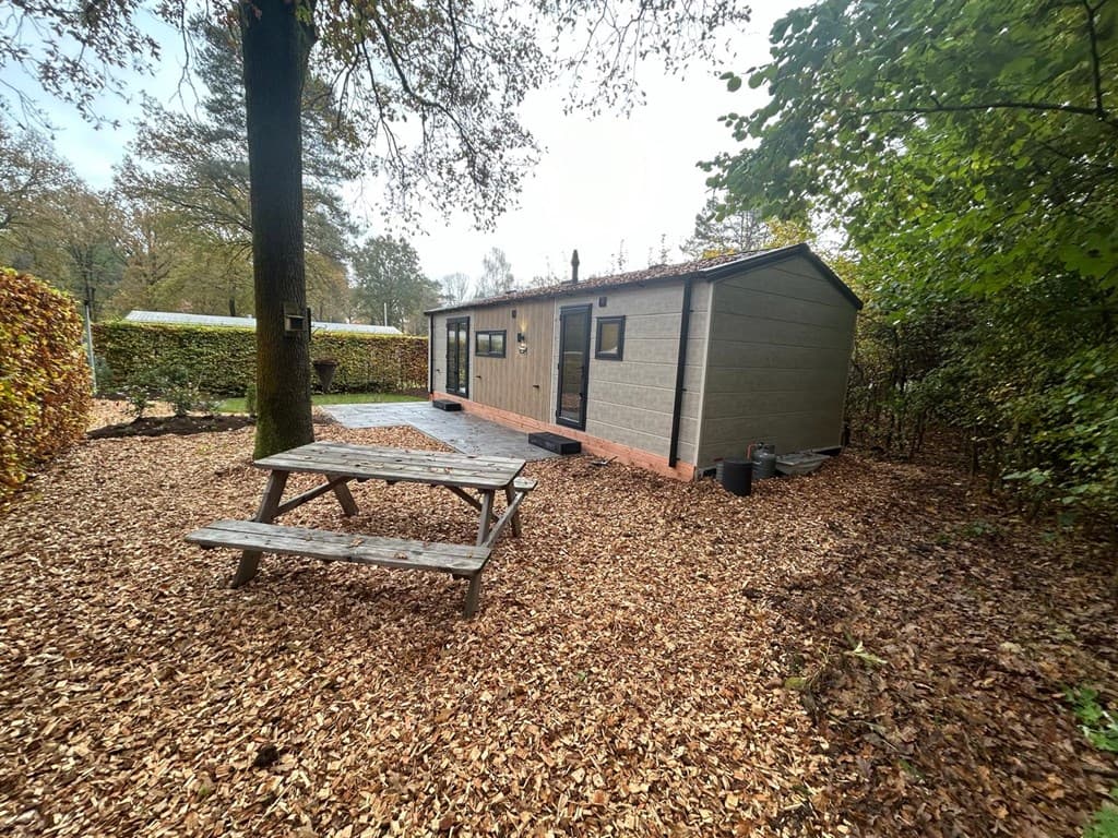 Vakantiehuisje omgeven door bladeren, met een houten picknicktafel en groene begroeiing aan de zijkanten.