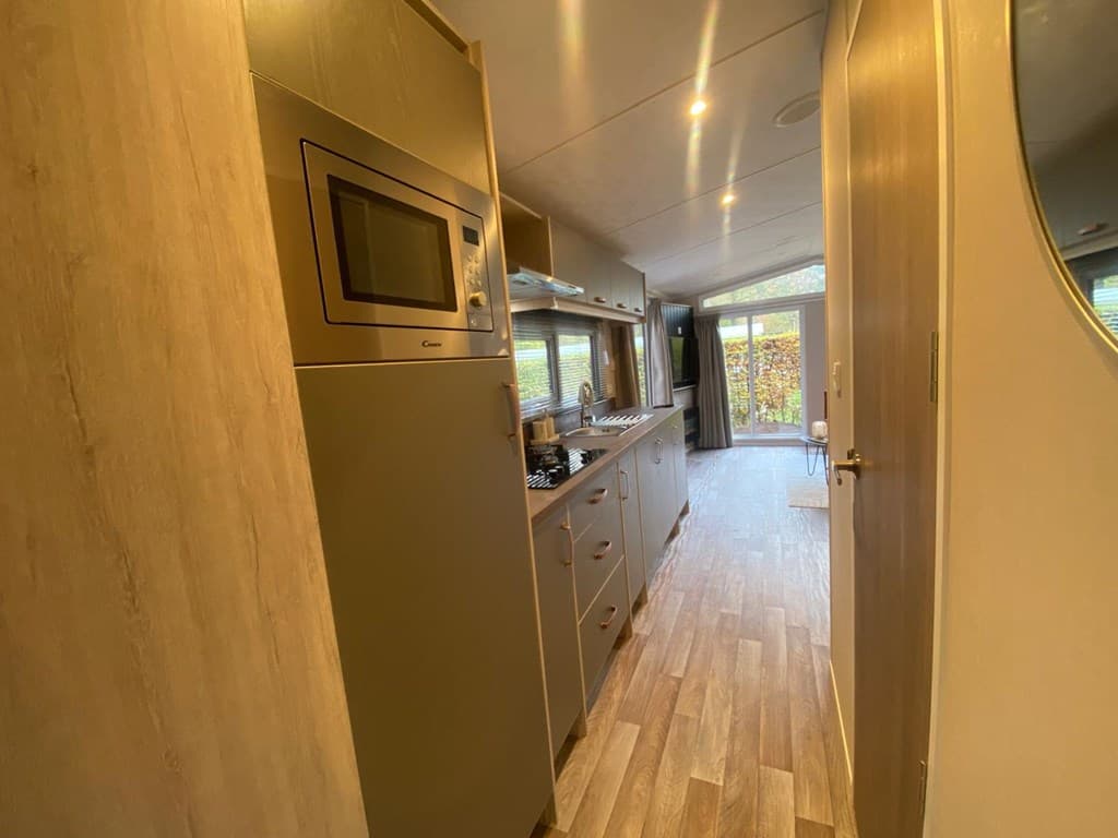 Inzicht in een moderne keuken met apparatuur en een doorgang naar een lichte woonruimte met ramen naar buiten.