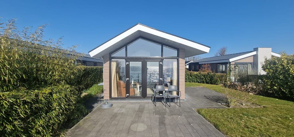 Moderne vakantiewoning met grote ramen, terras en uitzicht op het water, omgeven door groene hagen en gazon.