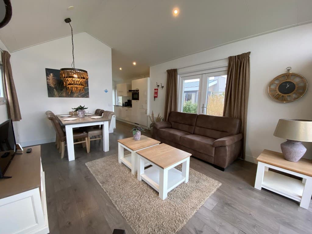 Woonkamer met een bruine bank, eettafel, tv en moderne inrichting, met grote ramen en zichtbaar keukengedeelte