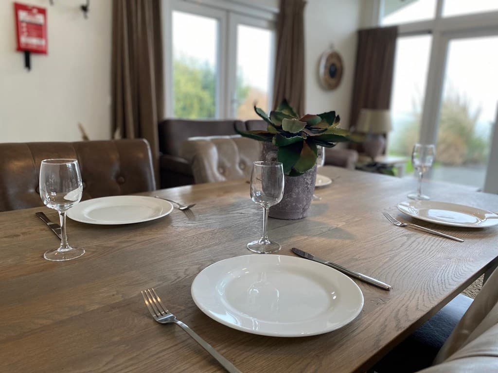 Eettafel met witte borden, glazen en een plant, met zicht op een lichte woonkamer en grote ramen.