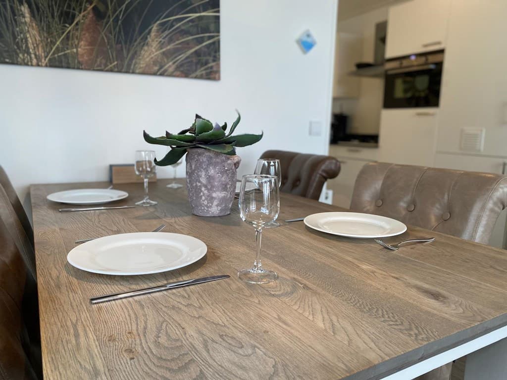 Eetkamertafel met plantaardig decor, borden en bestek, in een lichte ruimte met een zicht op de keuken.