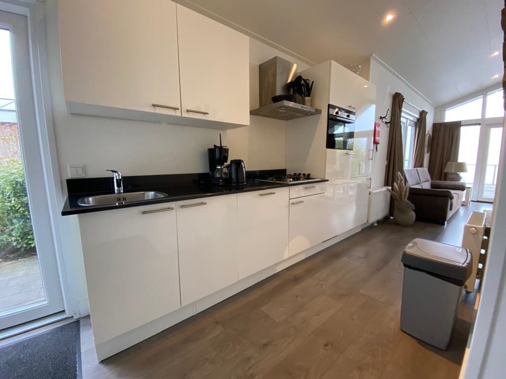 Moderne keuken met witte kasten, zwarte bladen en apparatuur, met uitzicht op een zithoek en ramen aan de rechterzijde.