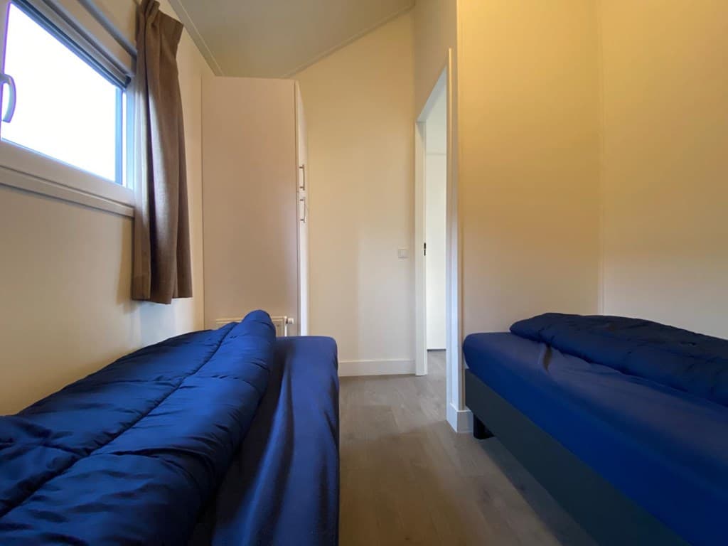 Twee eenpersoonsbedden met blauwe dekbedden in een lichte slaapkamer met ramen en kast.