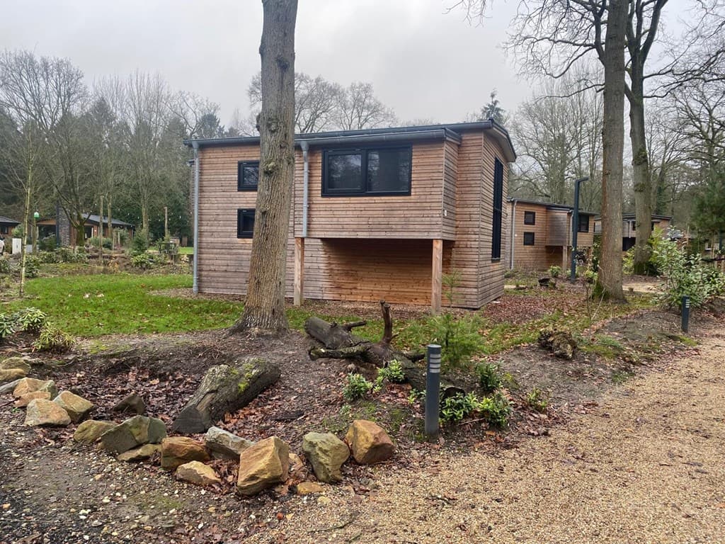 Houten vakantiehuis op een ruim perceel met bomen en een grindpad eromheen, omgeven door groene planten en struiken