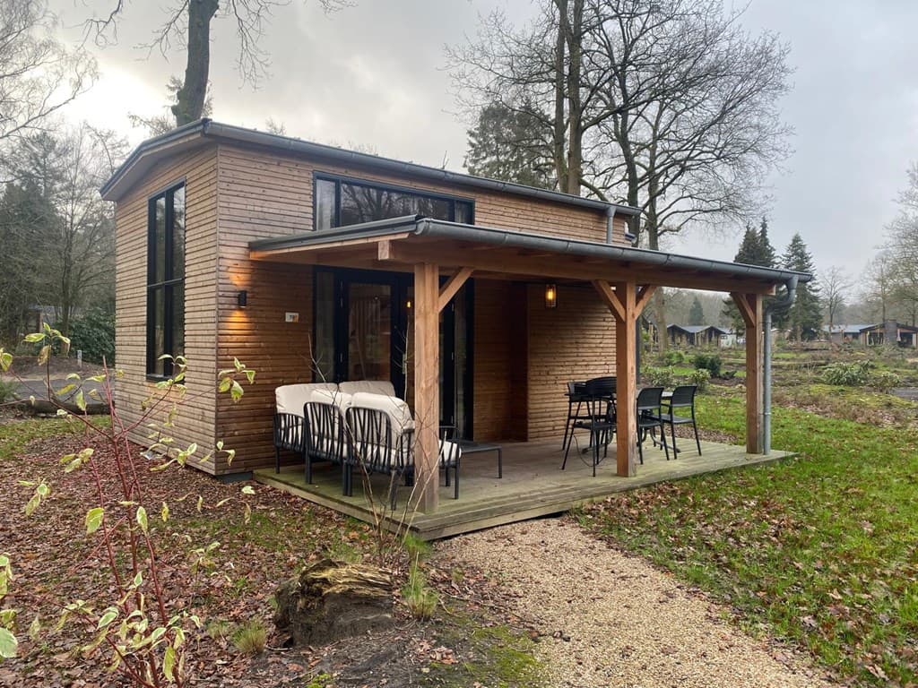 Houten vakantiehuis met overdekte veranda, grote ramen en een tuin met gras en bomen rondom.