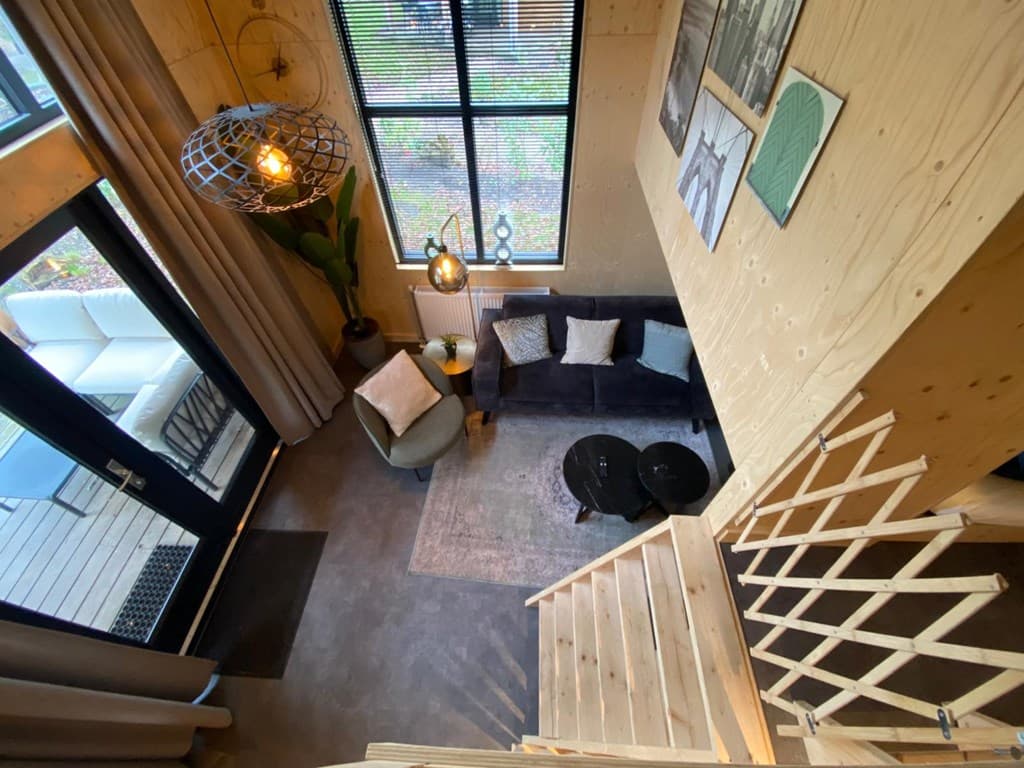 Zicht op de woonkamer met bank, stoelen, tafels en grote ramen met uitzicht op buitenruimte en trap naar boven.