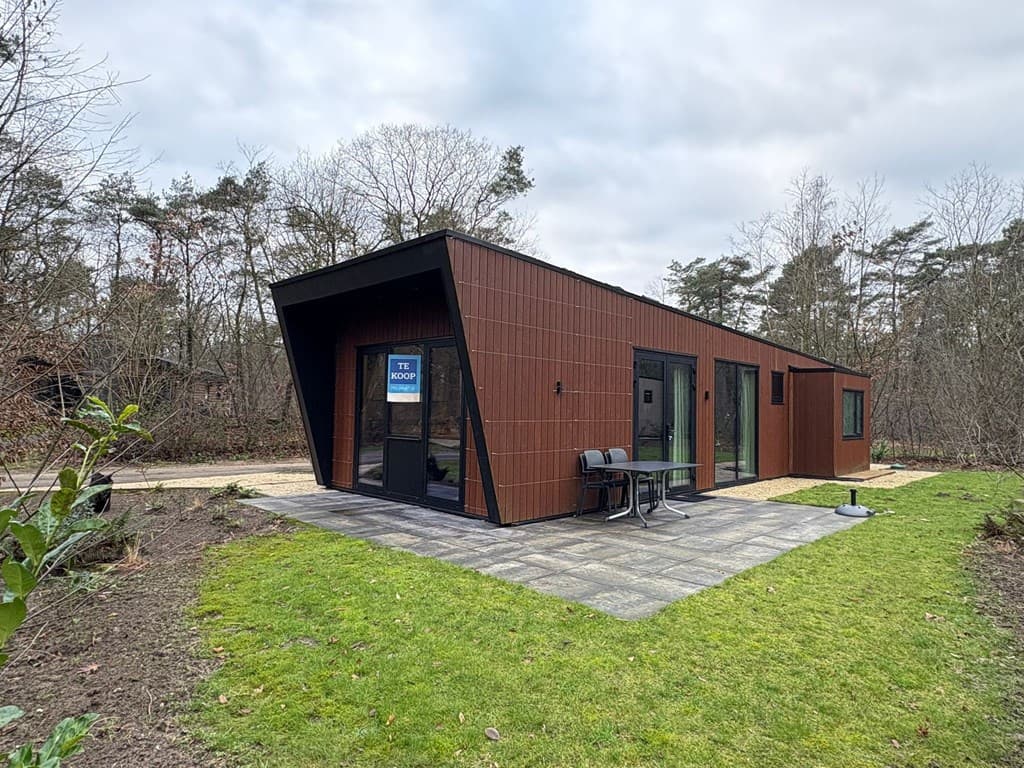 Moderne vakantiewoning met een houten gevel, grote ramen en een terras met stoelen, omgeven door bosrijk gebied.