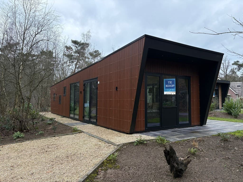 Moderne vakantiewoning met houten gevel en grote ramen, gelegen aan een gravelpad omringd door bomen en groen.