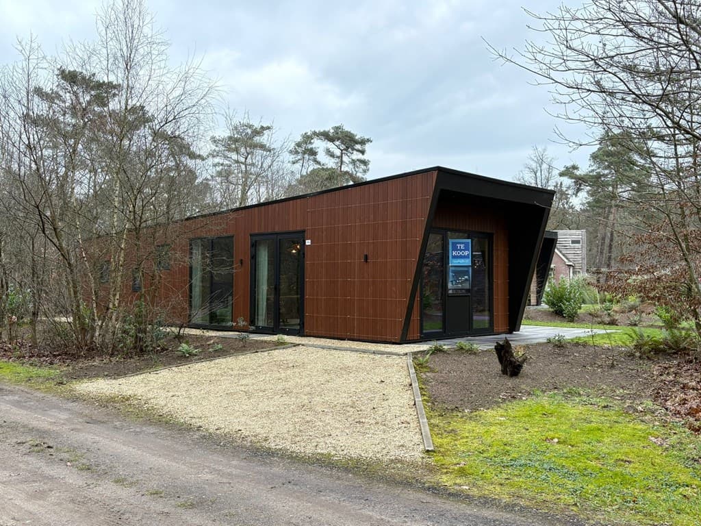 Moderne vakantiewoning met een hoekige gevel, omringd door bomen en een grindpad naar de voordeur