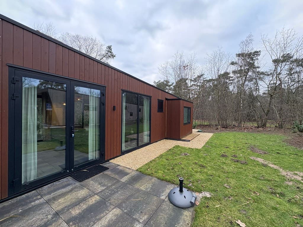 Moderne vakantiewoning met houten gevel, grote ramen en betegeld terras met gras en bomen op de achtergrond