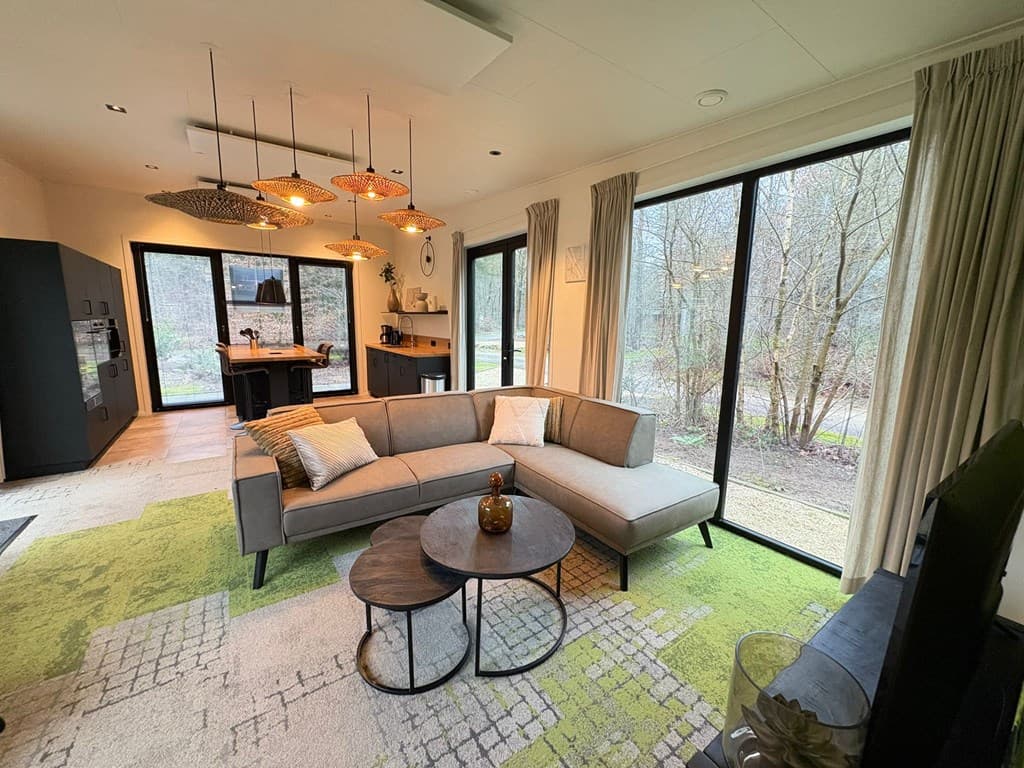Moderne woonkamer met grote ramen, loungebank, salontafel en open keuken met uitzicht op een groene omgeving