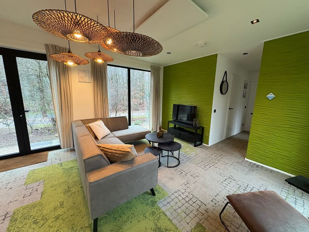 Woonkamer met groene wand, bank, tafel en TV, grote ramen met uitzicht op de natuur.