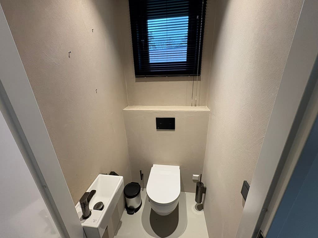 Toiletruimte met moderne toiletafwerking, venster met jaloezieën en minimalistische elementen.