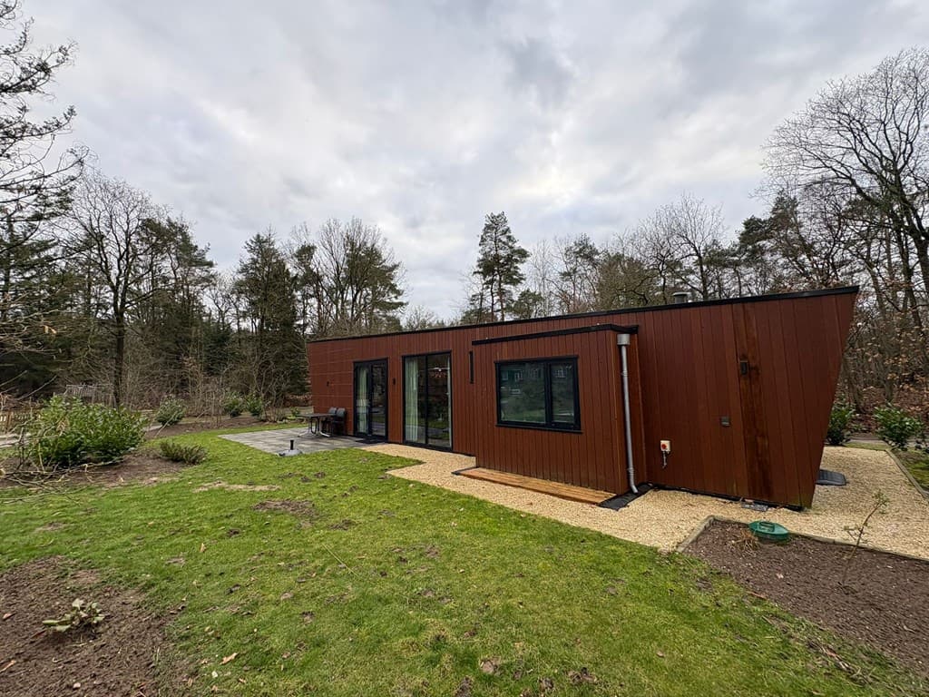 Moderne houten vakantiewoning met grote ramen en een aangelegde tuin, omgeven door bomen en gras.