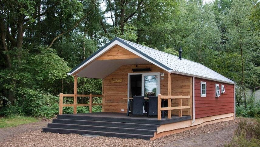Houten vakantiehuis met een overdekte veranda en een toegangstrap, omringd door bomen en groen.