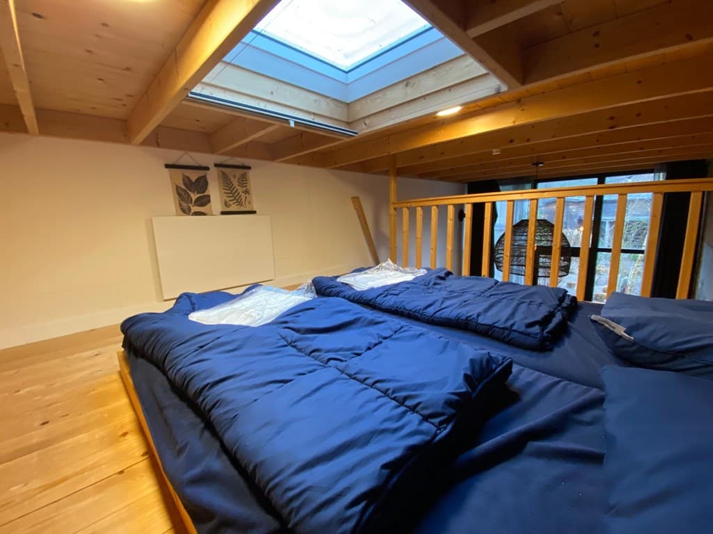 Zolderverdieping met houten balken, lichtinval vanuit een dakraam en twee opgemaakte bedden met blauwe dekbedden