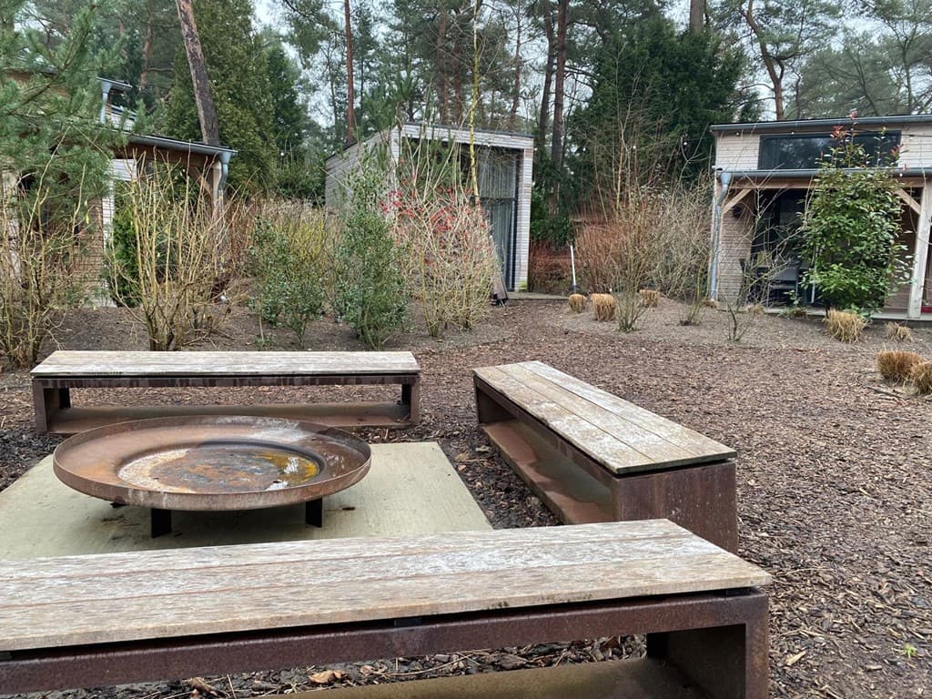 Tuin met zitbanken rond een vuurplaats, omringd door begroeiing en enkele moderne gebouwen in de achtergrond