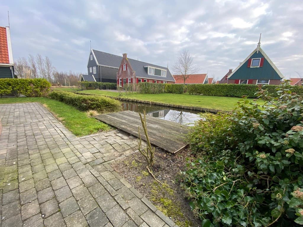 Tuinoppervlak met houten vlonder en waterpartij, omringd door gras en groen, met woningen op de achtergrond.