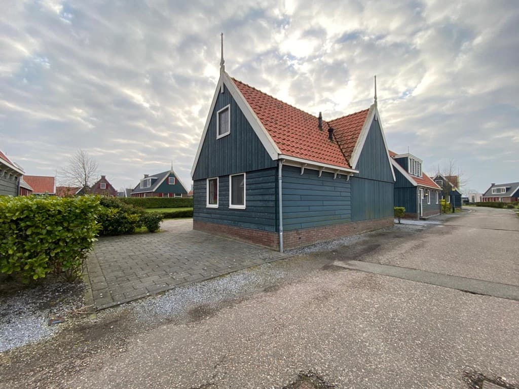 Houten vakantiehuis met een schilddak en rode pannen, gelegen aan een verhard pad met groenheken.