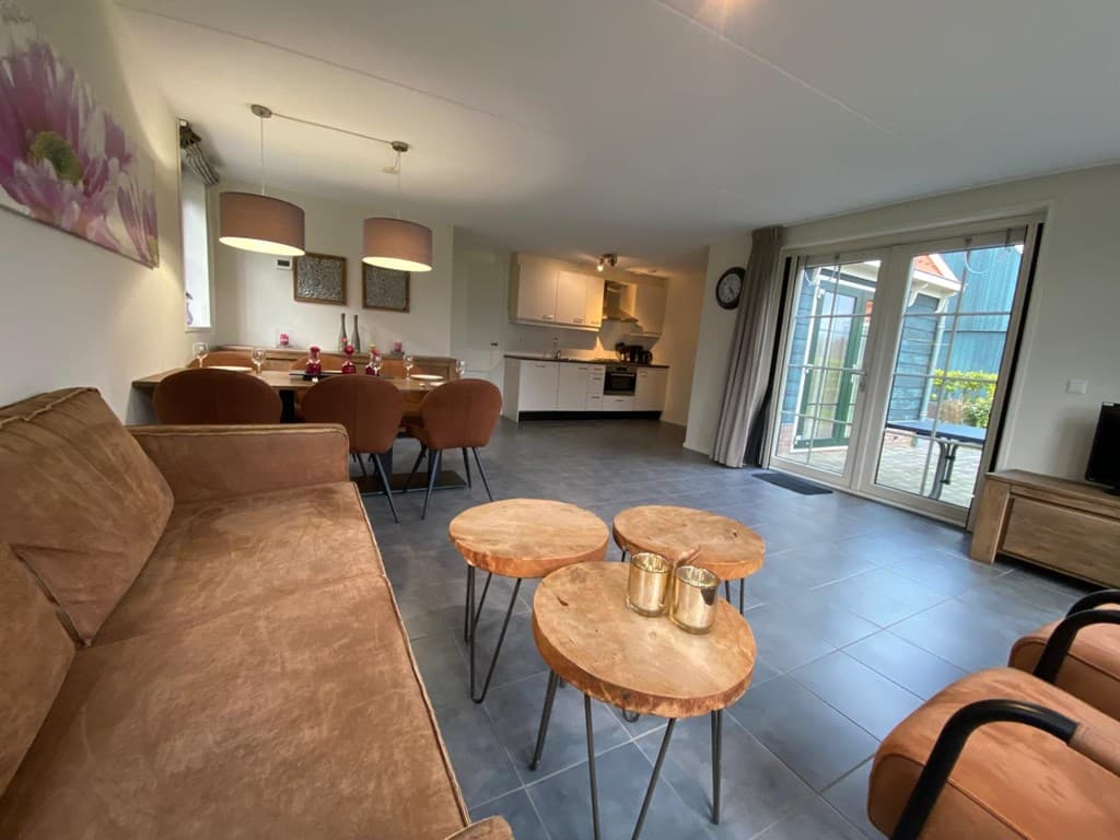 Woonkamer met een bank, houten tafels, eethoek en toegang tot een terras met glazen deuren