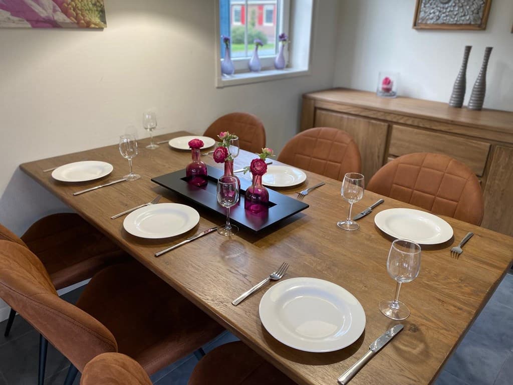 Eetkamertafel met opgemaakte borden en glazen, decoratie met bloemen en een kast op de achtergrond.