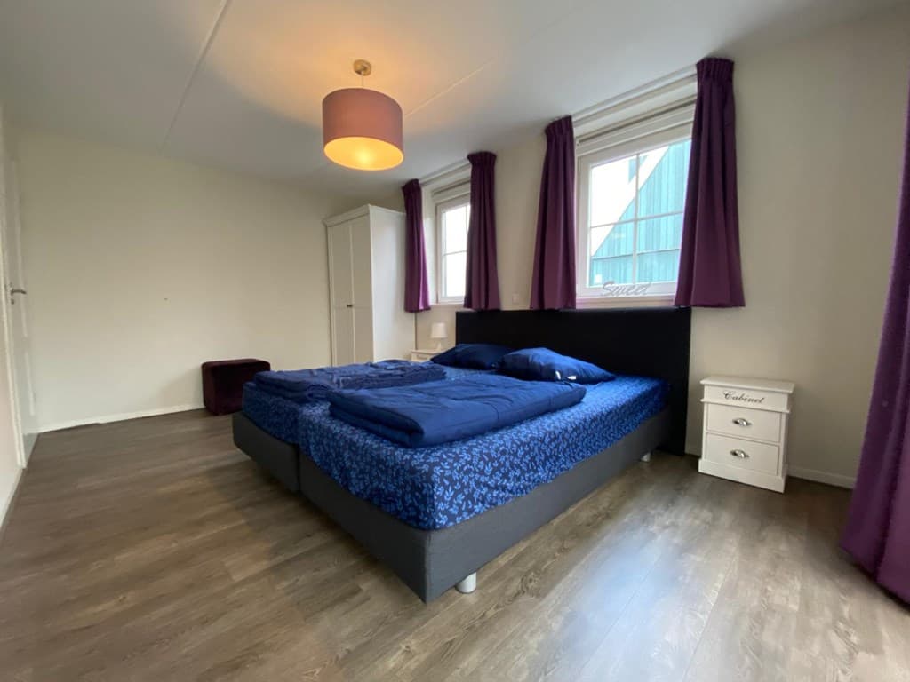 Slaapkamer met tweepersoonsbed, nachtkastje en kast; paarse gordijnen bij de ramen en laminaatvloer.