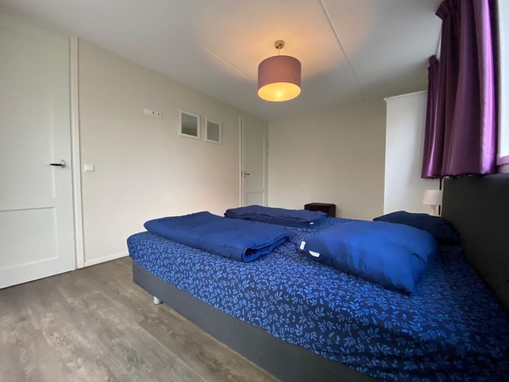 Slaapkamer met een tweepersoonsbed, paarse lamp en vitrage aan het raam, moderne vloer en een deur naar een andere ruimte