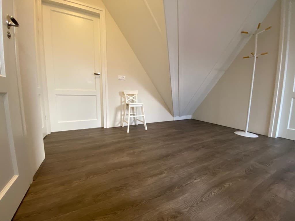 Lichte kamer met een houten vloer, een witte stoel en een klemmende deur onder een schuine wand