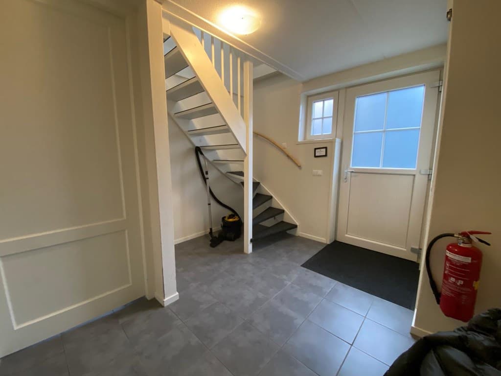 Hal met betegelde vloer, trap naar boven en een buitendeur met bovenraam, brandblusser aan de wand en stofzuiger op de vloer.