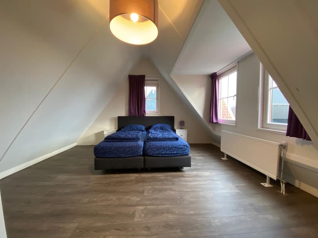 Slaapkamer met twee eenpersoonsbedden, houten vloer, grote ramen en paarse gordijnen in een schuin dak.