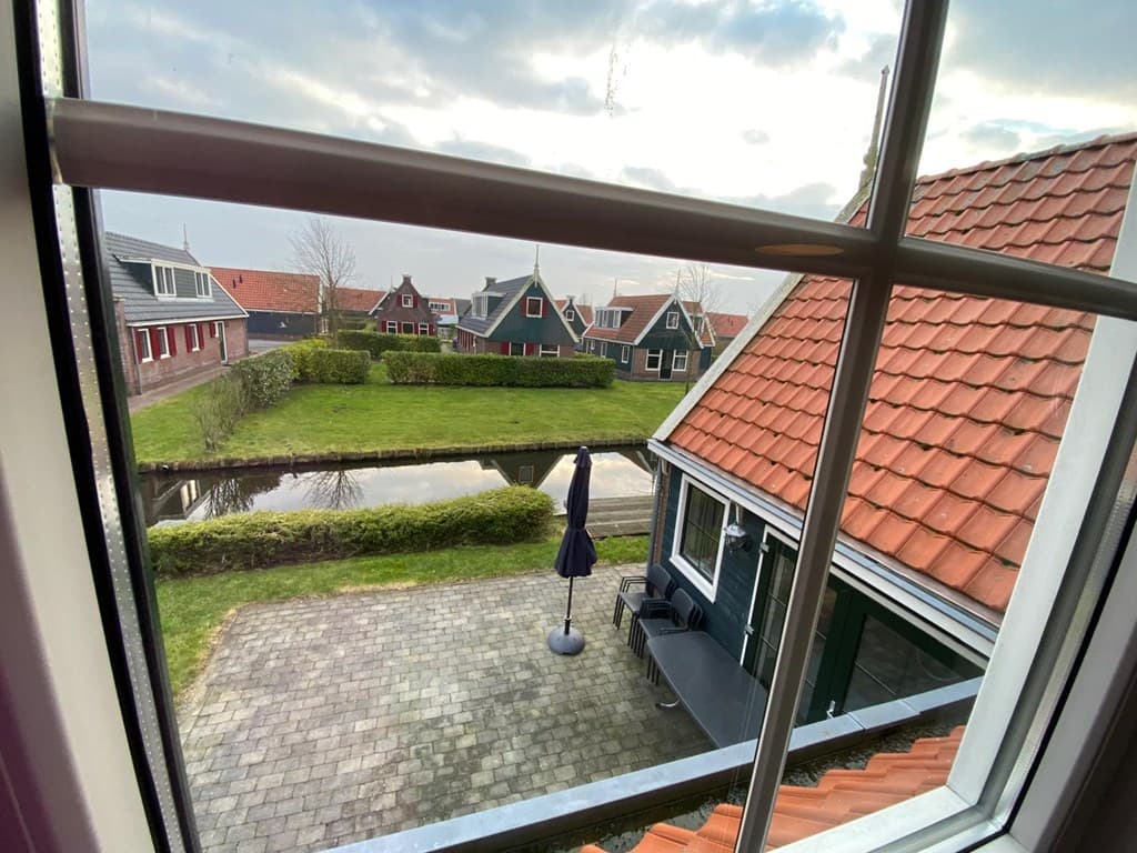 Uitzicht vanuit een raam over een betegelde tuin met een terras en parasol, tegenover watervijvers en groene hagen