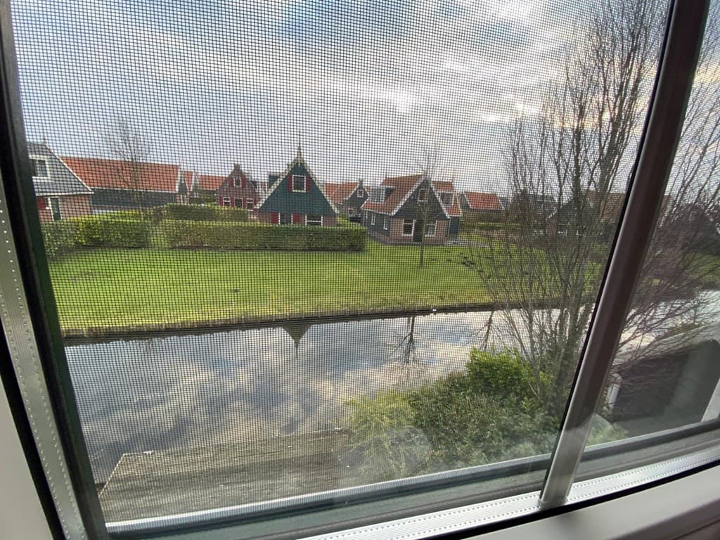 Uitzicht vanuit een raam op een tuin met gras en een sloot, omringd door woningen met typisch Nederlandse daken.