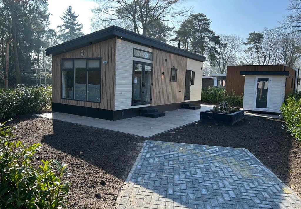Moderne vakantiewoning met houten gevel en groot terras, omgeven door groen en een verhard pad.