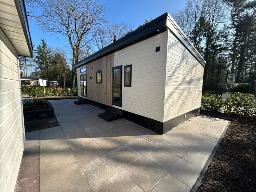 Moderne vakantiewoning met een lichte gevel en verhard terras, omgeven door bomen en groene struiken.