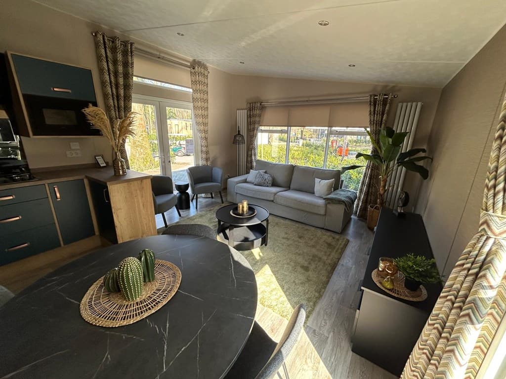 Woonkamer met een bank, stoelen, een ronde tafel en planten met grote ramen voor natuurlijk licht.