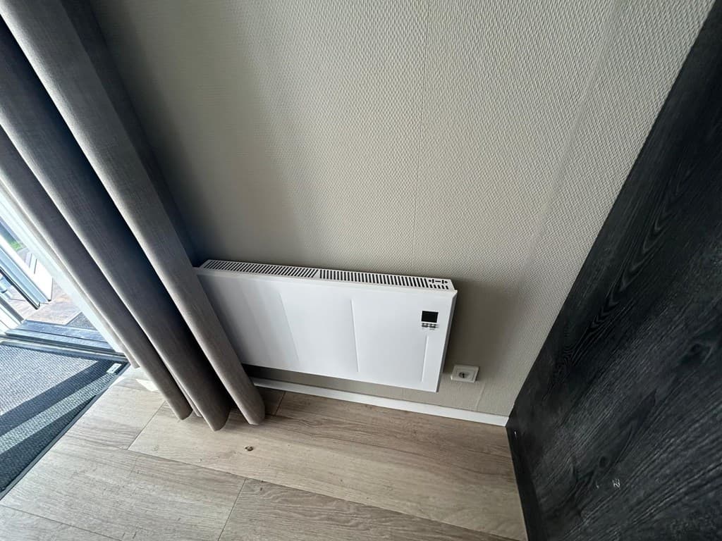 Een witte verwarmingseenheid tegen een muur met beige wand en houten vloerbedekking.