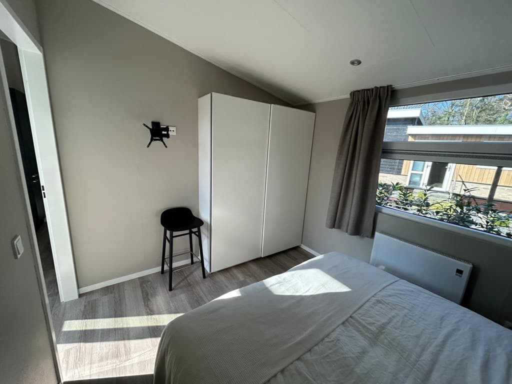 Slaapkamer met een bed, witte kast, stoel en raam met uitzicht op de buitenruimte.