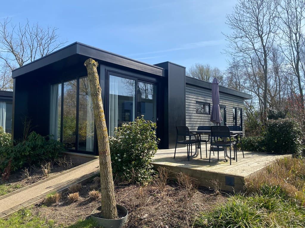 Moderne vakantiewoning met houten terras en tuinstoelen, omringd door groen en bomen.